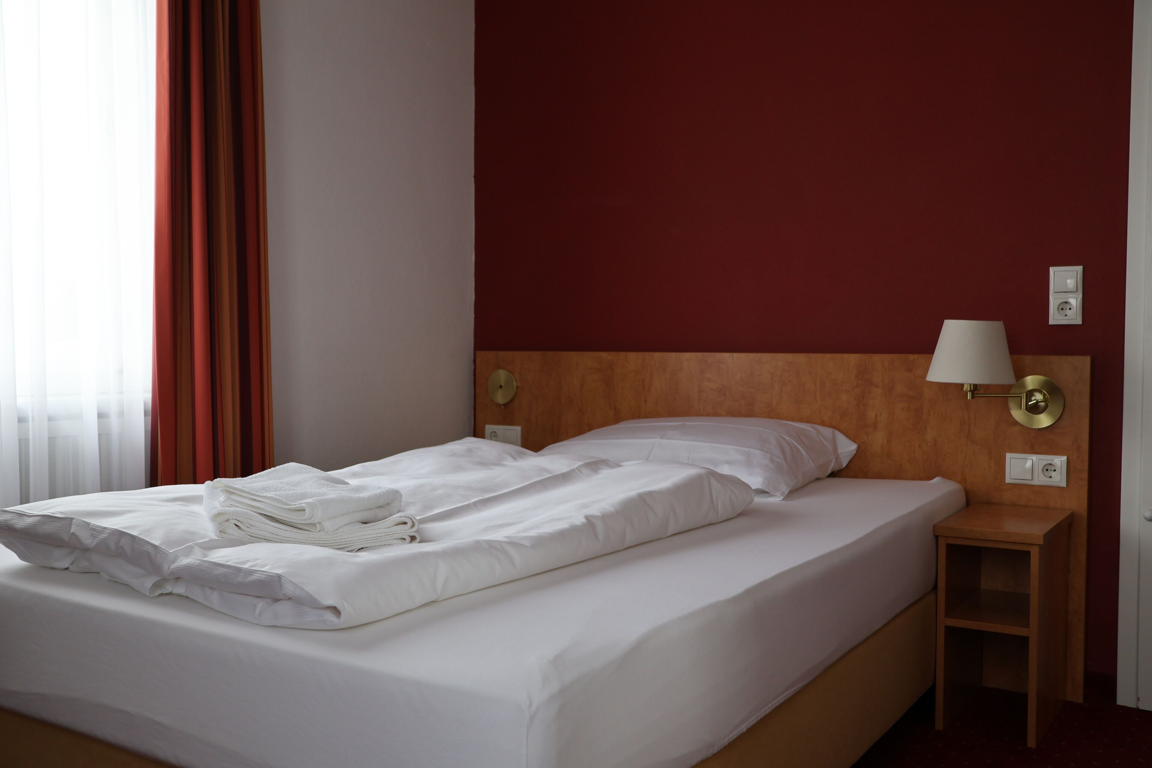 A hotel room with a double bed, a wooden headboard, a nightstand with a lamp, and a red accent wall.