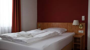 A hotel room with a double bed, a wooden headboard, a nightstand with a lamp, and a red accent wall.