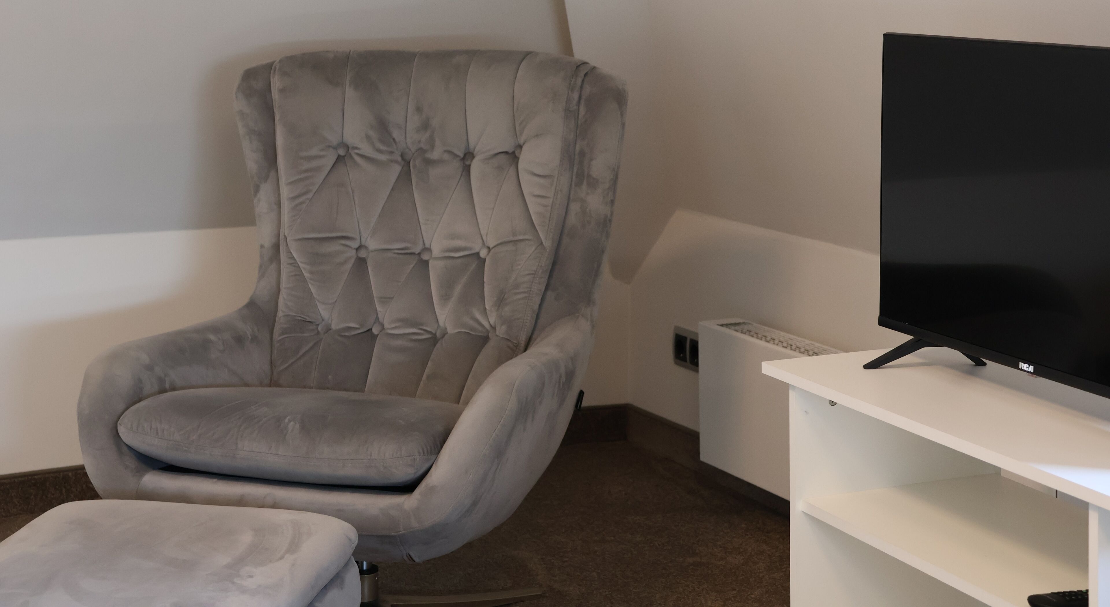Un sillón gris con capitoné y un reposapiés, un televisor de pantalla plana sobre un soporte blanco y un mueble de entretenimiento blanco.