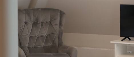 Una habitación con un sillón gris con capitoné, un pequeño reposapiés, un mueble blanco para televisión y un radiador.