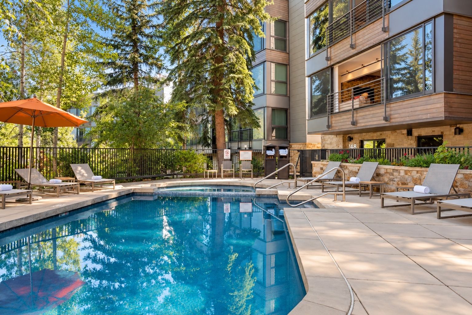 Une piscine dotée de chaises longues et d’une terrasse en bois, entourée d’arbres et d’un bâtiment aux grandes fenêtres.