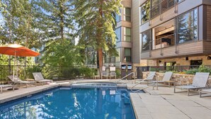 Une piscine dotée de chaises longues et d’un parasol, entourée d’arbres et bordée d’un bâtiment moderne.