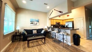 A living room with a sofa, coffee table, and a kitchen area with a bar and counter. - Dances with Waves - Villagio C245 Condo in Beautiful Perdido Key, FL (Pensacola)