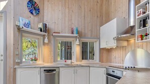 Une cuisine moderne avec des meubles blancs, un lave-vaisselle, un four et un barbecue intégré. On y trouve des fenêtres avec des rideaux, une horloge et une étagère avec des livres et des objets décoratifs.