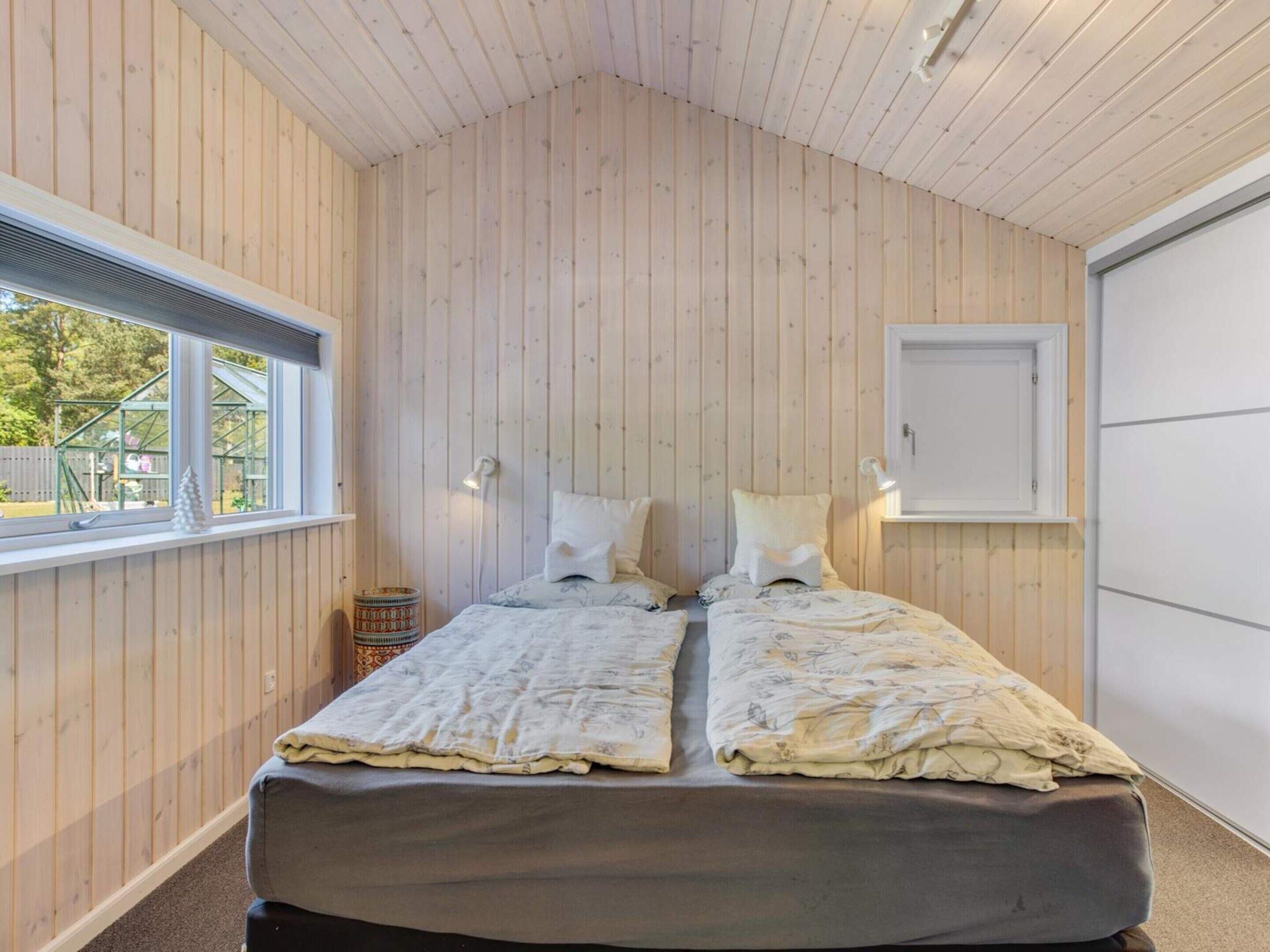 A room with wooden panel walls, a bed with two pillows, a window with a view of greenery, and a door.