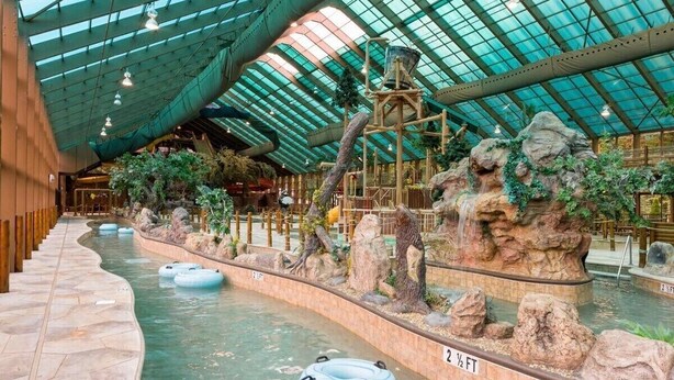 Parque acuático interior con techo de cristal, formaciones rocosas artificiales y flotadores inflables.