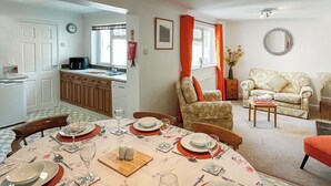 A dining area with a table set for six, a kitchen in the background, and a living room with a sofa and armchair. - Escape to Barnhayes, a charming retreat in the picturesque village of Hartland. (Hartland)