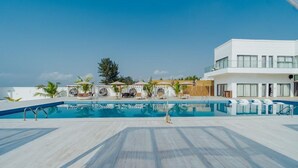 Area piscina sul tetto con sedie a sdraio e ombrelloni, un edificio moderno con ampie finestre e cielo azzurro.