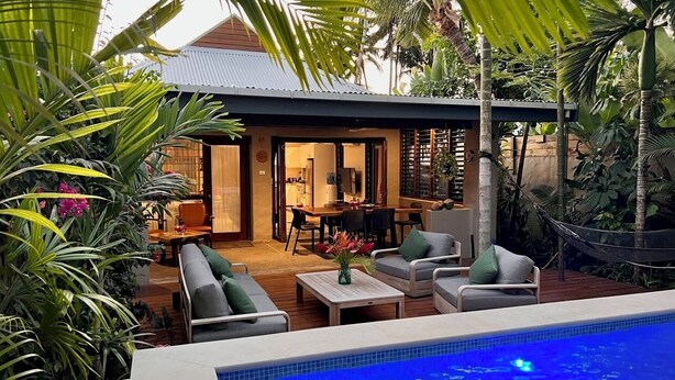 Zona junto a la piscina con mobiliario exterior, comedor y exuberante vegetación tropical.