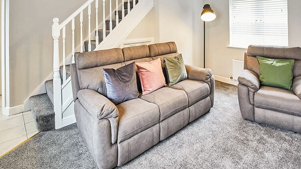 Un salon avec un canapé beige, un fauteuil marron, un escalier et une grande fenêtre avec des stores.