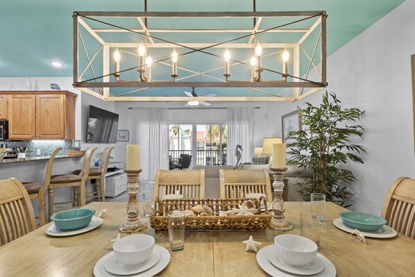 A dining area with a wooden table set for four, surrounded by wicker chairs. A decorative basket is placed in the center, and the room features a ceiling fan and a hanging light fixture.