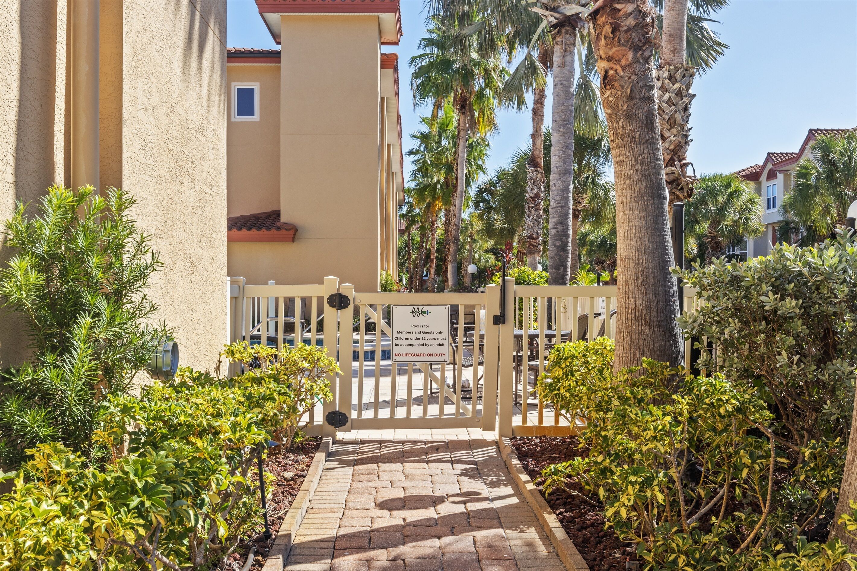 A gated entrance to a building with a paved pathway, surrounded by lush greenery and palm trees.