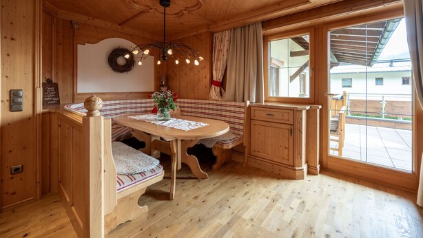 A wooden cabin interior with a round dining table, bench seating, and a large window offering a view of the outside. - Spacious vacation apartment in Kaiserwinkl - perfect for the whole family (Kössen)