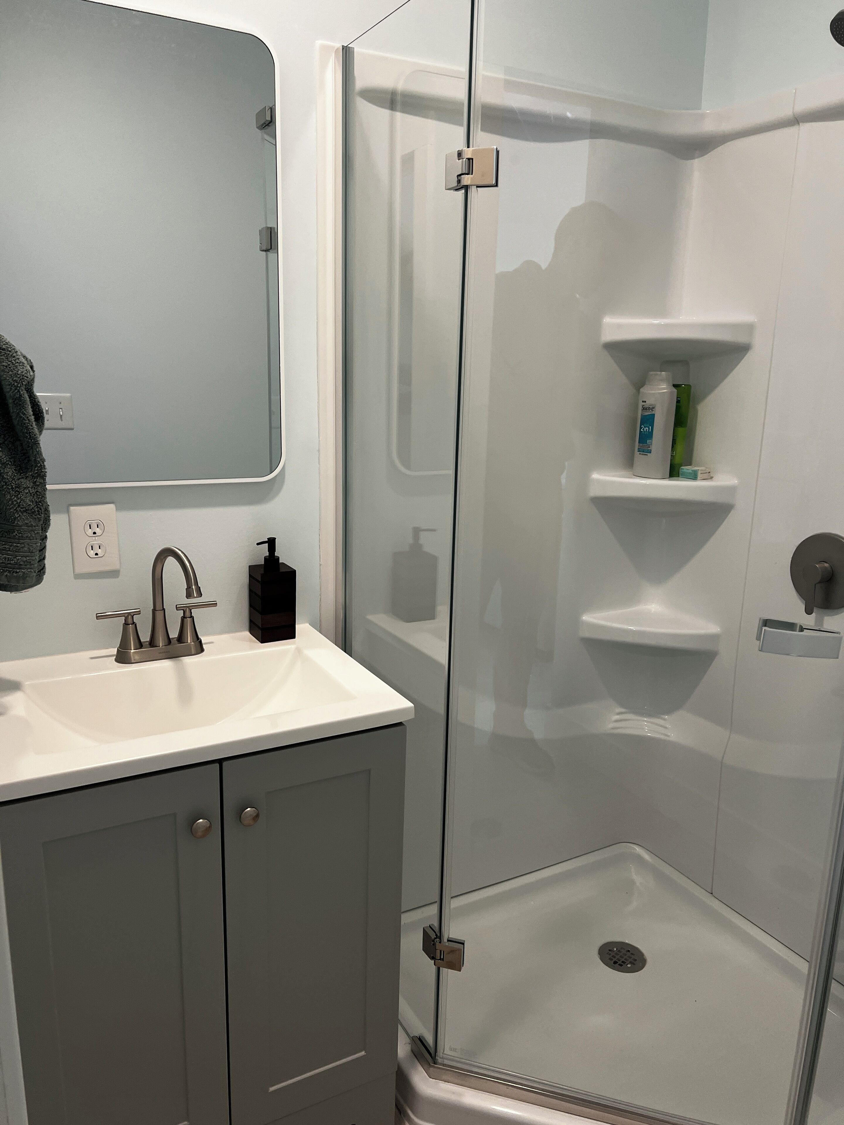 A bathroom with a white sink, a mirror, a glass shower enclosure, and a shelf with toiletries.