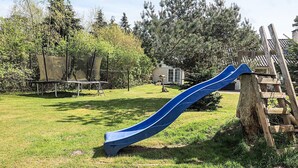 Een speeltuin met een blauwe glijbaan en een houten klimconstructie in een grasrijke omgeving met bomen en een huis op de achtergrond.