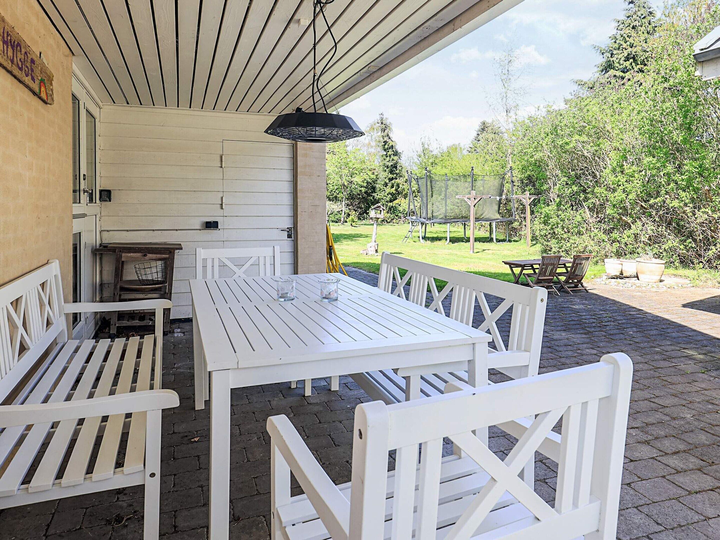 Un patio al aire libre con mobiliario de madera blanca, incluyendo una mesa y bancos, bajo un área cubierta con una lámpara colgante.