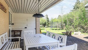 Een terras buiten met witte houten meubels, waaronder een tafel en bankjes, onder een overdekt gedeelte met een hanglamp.