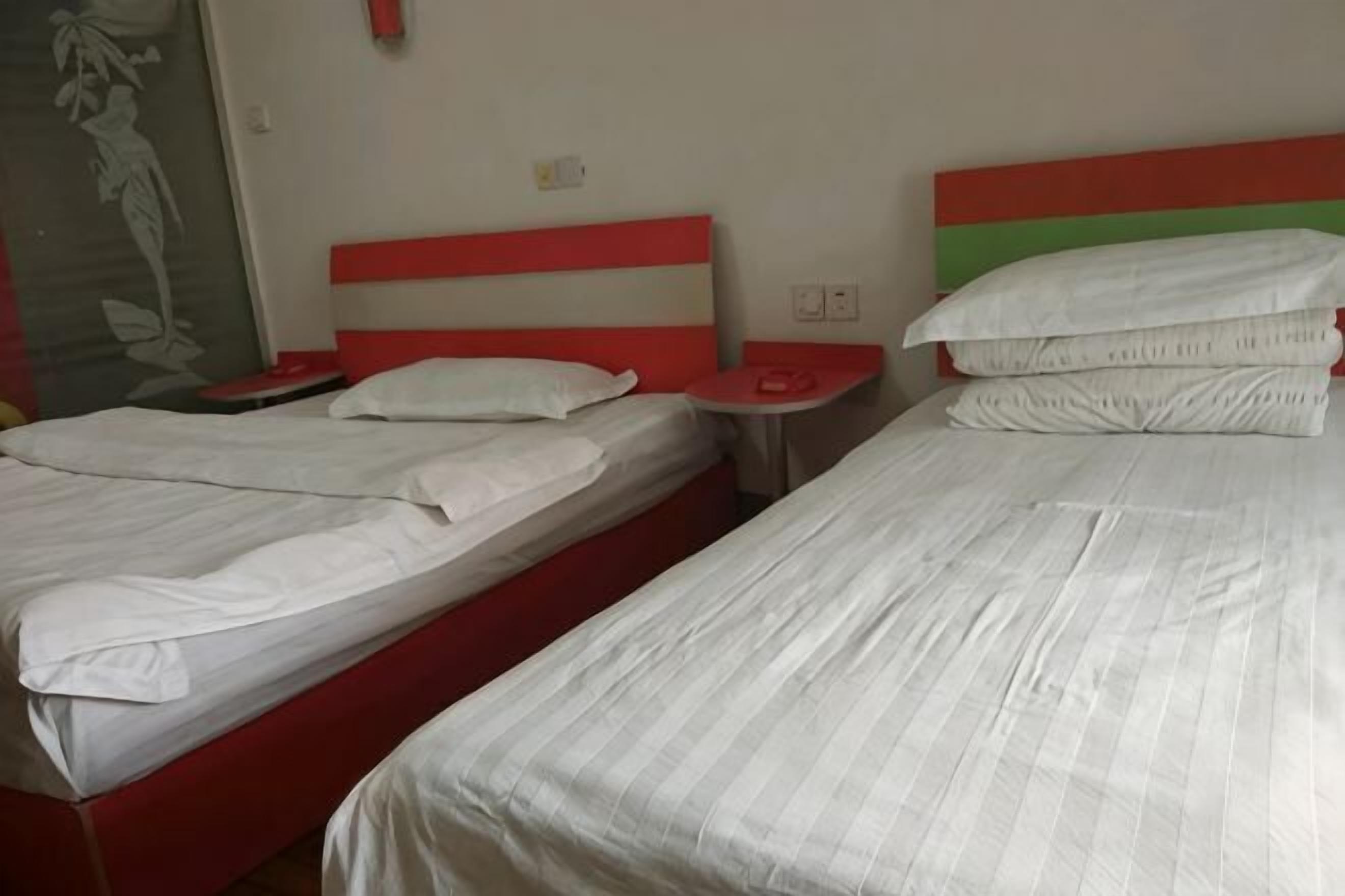 Hotellihuoneessa on kaksi sänkyä, joissa on valkoiset vuodevaatteet ja punaiset päätyosat. Sängyissä on yöpöydät, joissa on lamput, sekä pieni seinälle kiinnitetty hylly.
