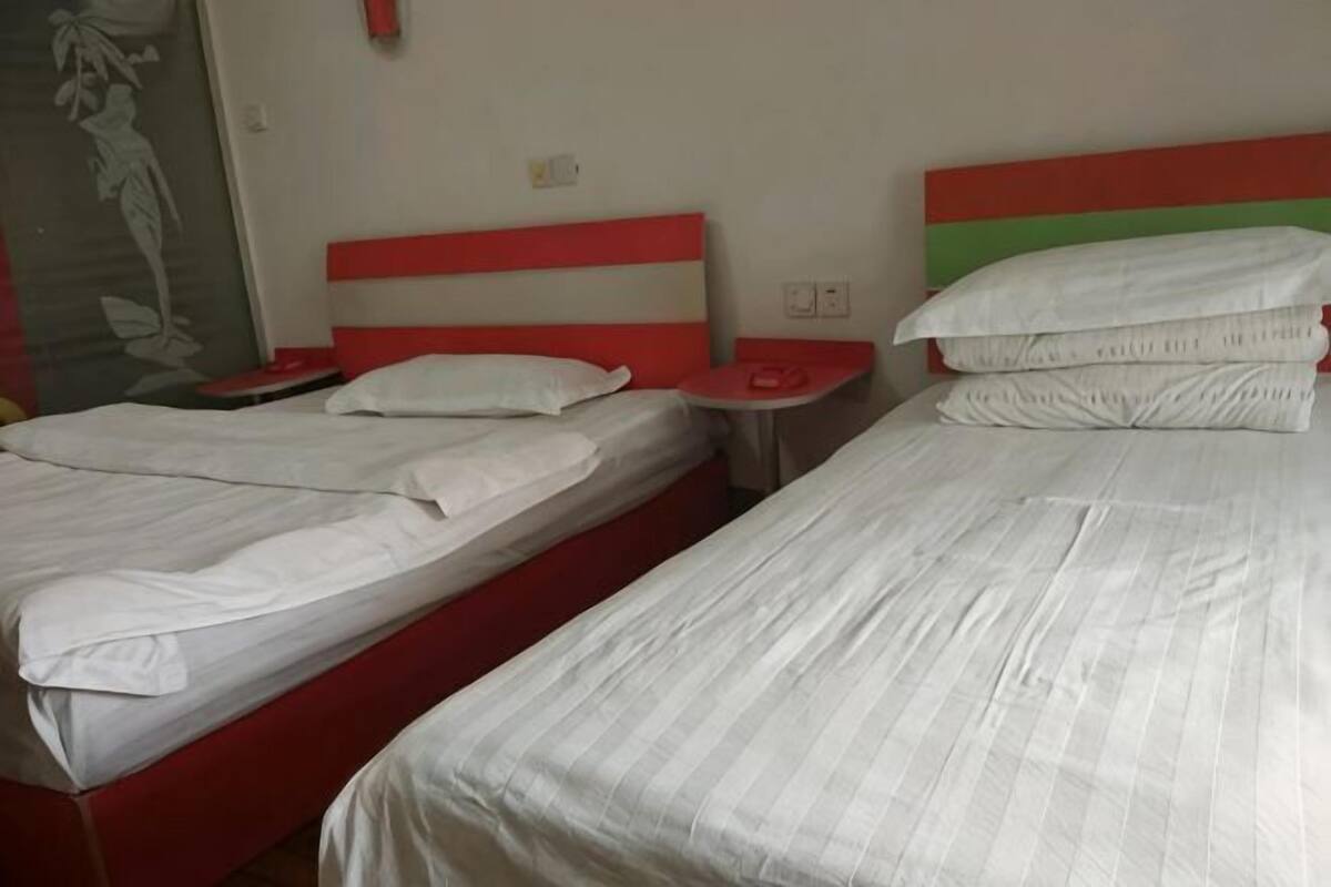 A hotel room with two beds, each with white bedding and red headboards. There are bedside tables with lamps and a small wall-mounted shelf.