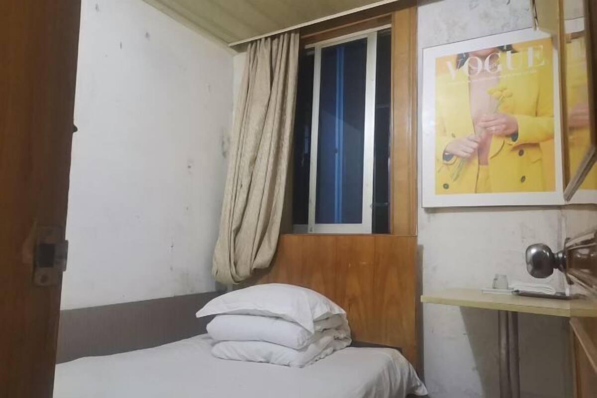 A bedroom with a bed, a window with a curtain, a wooden headboard, a desk with a lamp, and a magazine on the wall.