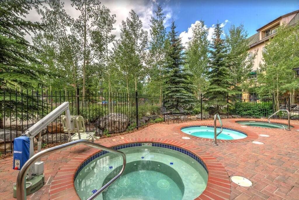 A pool area with two hot tubs surrounded by a brick patio, a metal fence, and trees in the background.