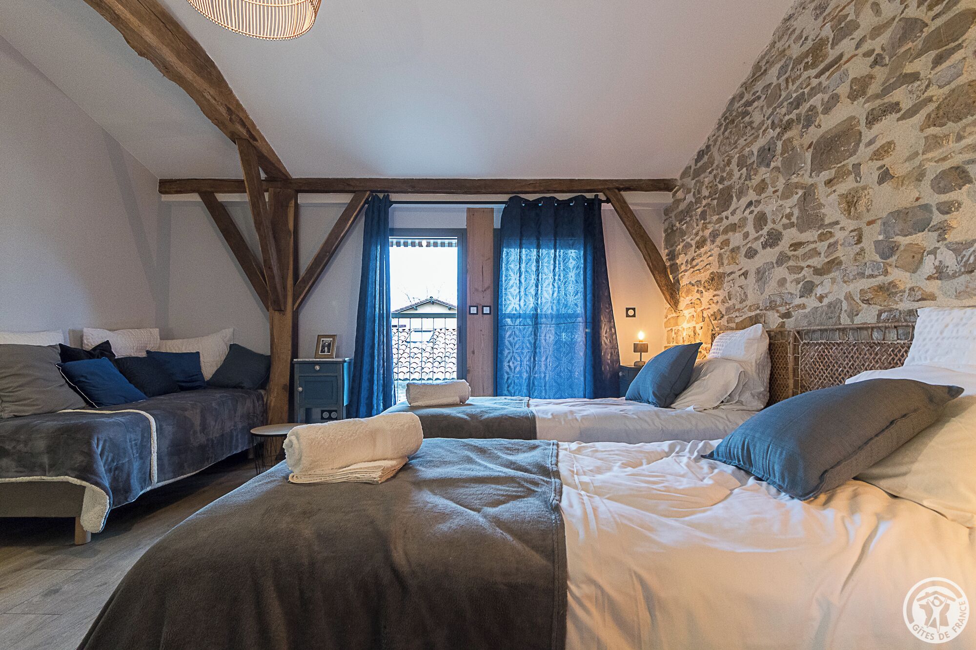 Ein Schlafzimmer mit zwei Betten, einer Steinwand, einer Holzbalkendecke und einem Fenster mit Vorhängen.
