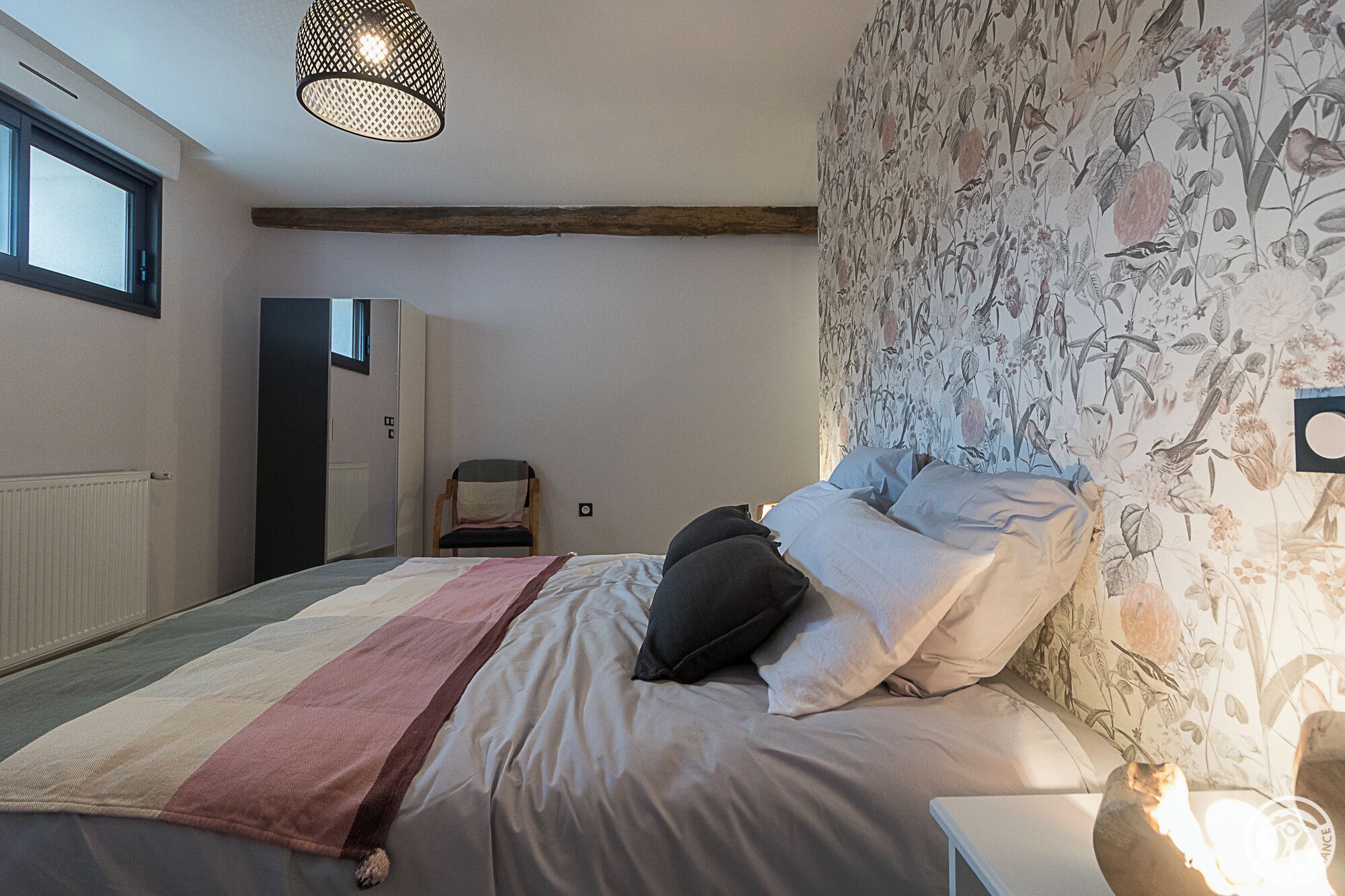 Un dormitorio con una cama, una silla, un radiador, un ventanal y una pared con papel tapiz floral.