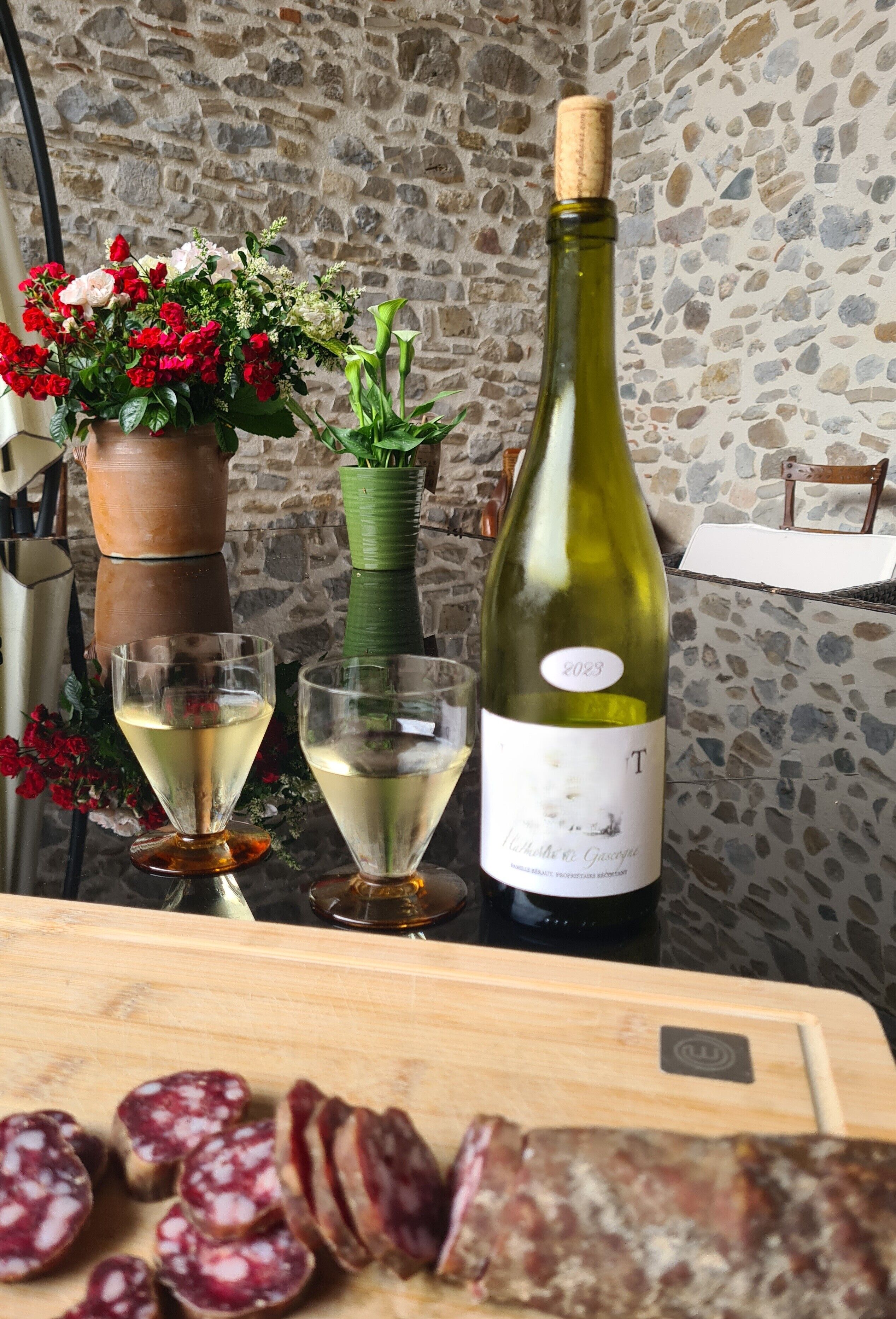A table with a bottle of wine, two glasses, a vase with flowers, and sliced salami.