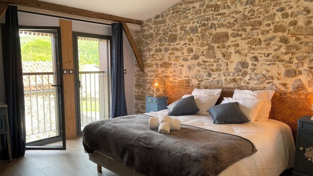 Un dormitorio con pared de piedra, una cama con almohadas, una mesita de noche y vistas a un espacio exterior a través de un vidrio.