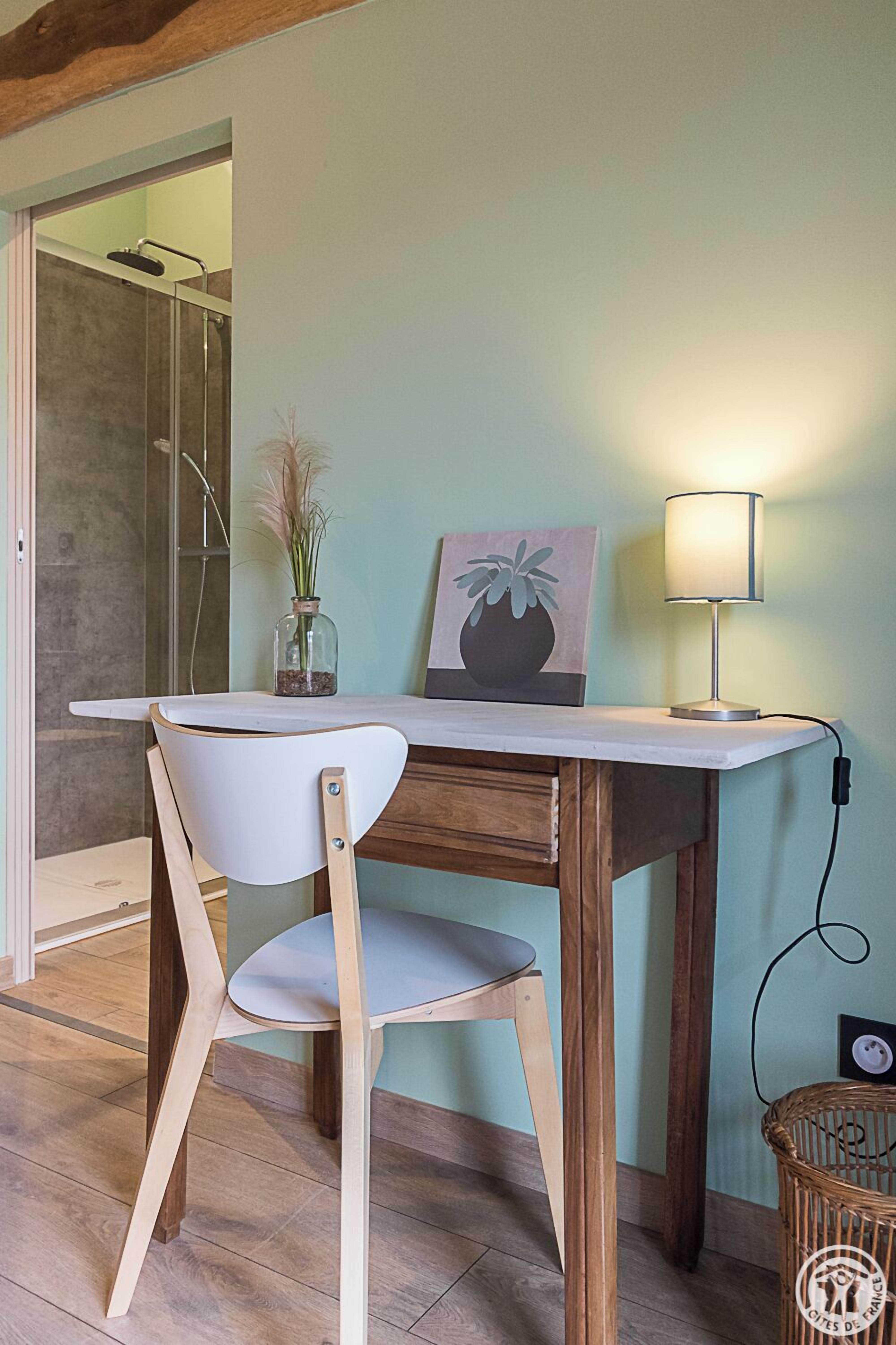 A room with a wooden desk, a white chair, a vase with dried plants, a lamp, and a wicker basket.