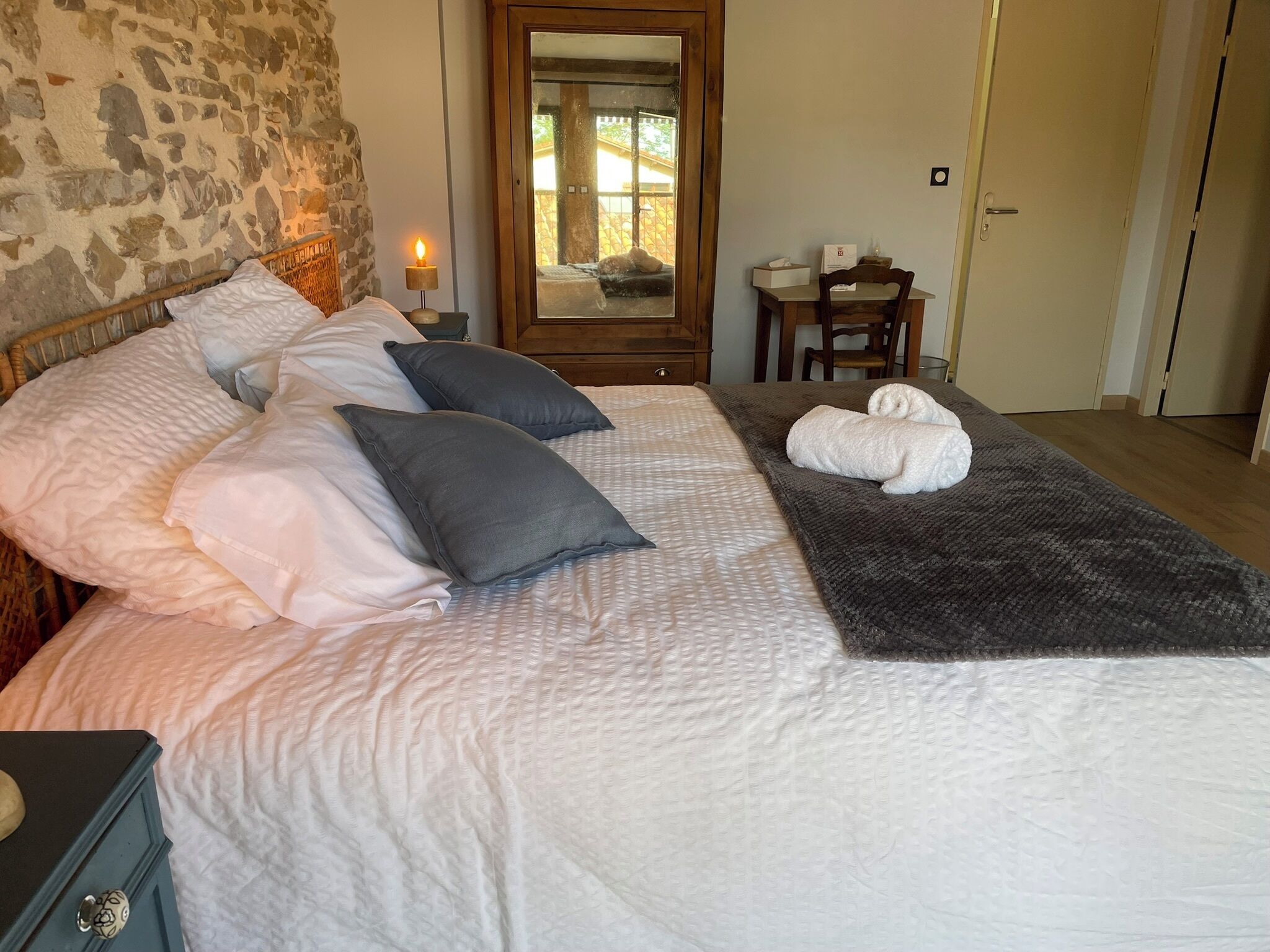 Un lit bien fait, avec une tête de lit, des oreillers et une couverture. On y trouve une table de chevet en bois avec une lampe, un miroir et une petite table accompagnée d’une chaise.