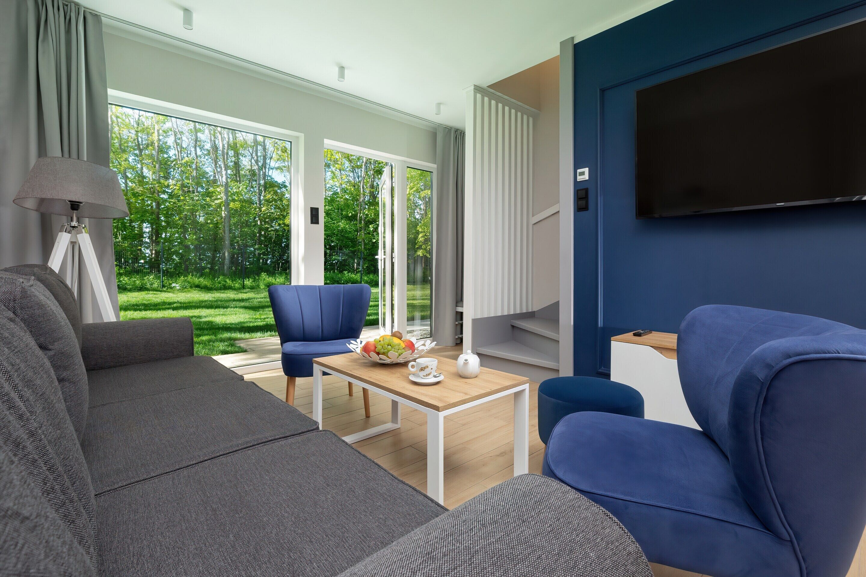 A modern living room with a sofa, armchair, and coffee table. There is a television mounted on the wall and a large window with a view of greenery outside.