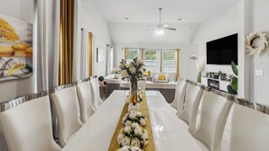 A dining area with a long table, white chairs, and a gold runner. There is a ceiling fan, a TV on the wall, and a fireplace. - Luxury 4BRS-3Baths Home, w/ King Bed & Private Gym (Katy)