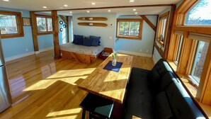 Une pièce spacieuse avec un plancher en bois, un lit avec une literie bleue, une table à manger agrémentée d’un vase et de grandes fenêtres.