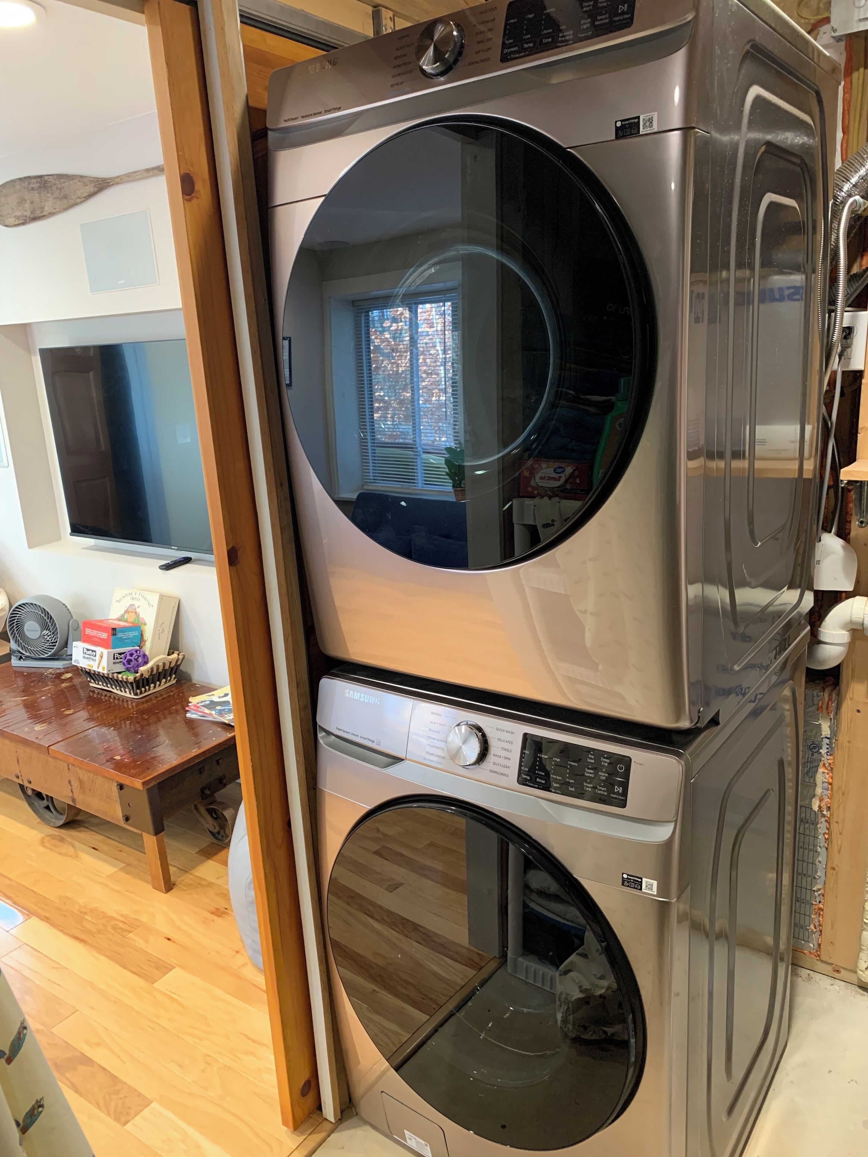 A stacked washer and dryer unit in a room with a wooden floor, a television, and a window with blinds.