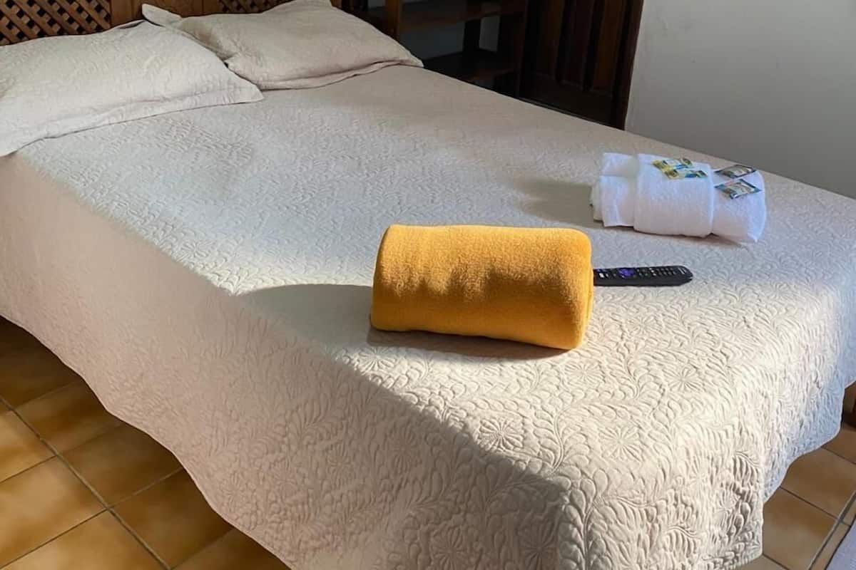 Una cama bien hecha con colcha blanca, cabecera de madera y un cuadro de un paisaje en la pared.