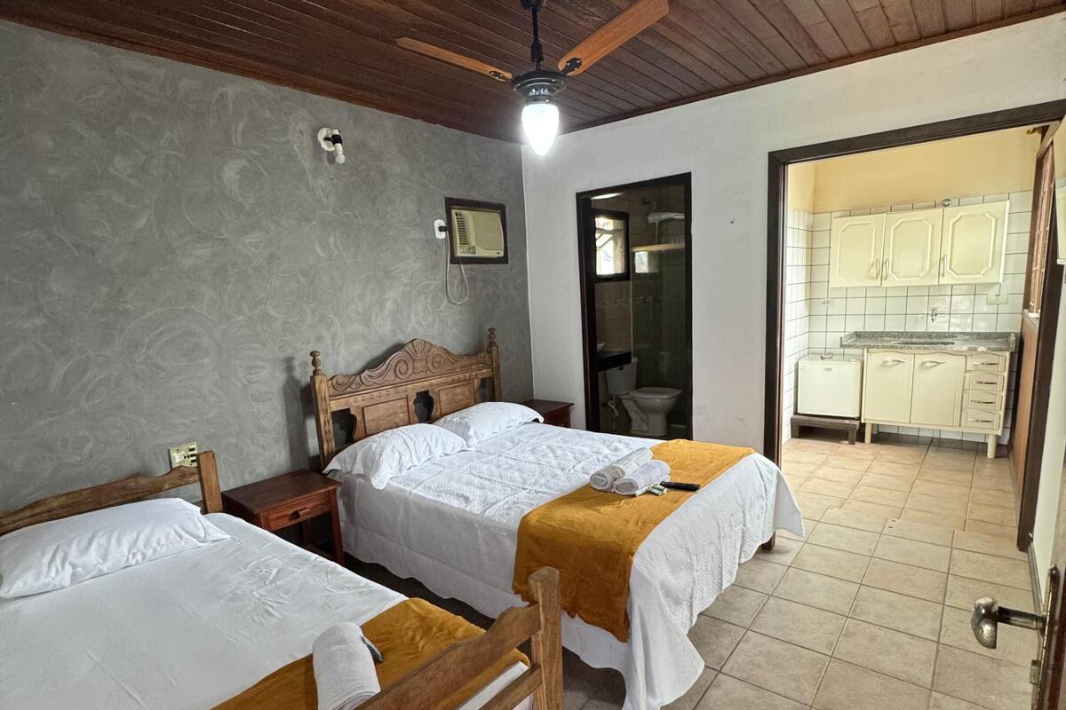 Una habitación con dos camas de madera, un ventilador de techo, una mesita y un baño visible a través de una puerta abierta.