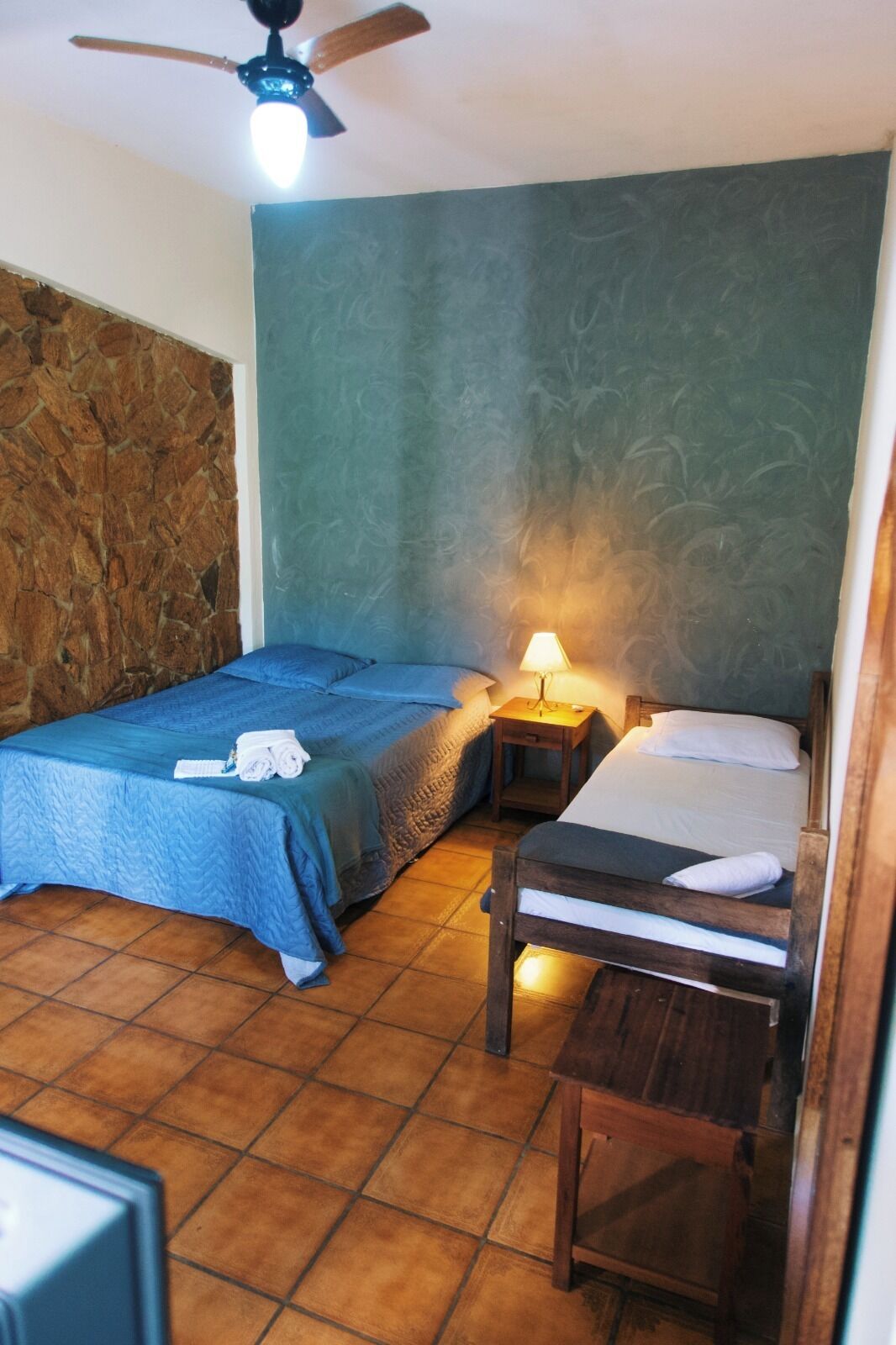 A room with two beds, a stone wall, a ceiling fan, and a tiled floor.