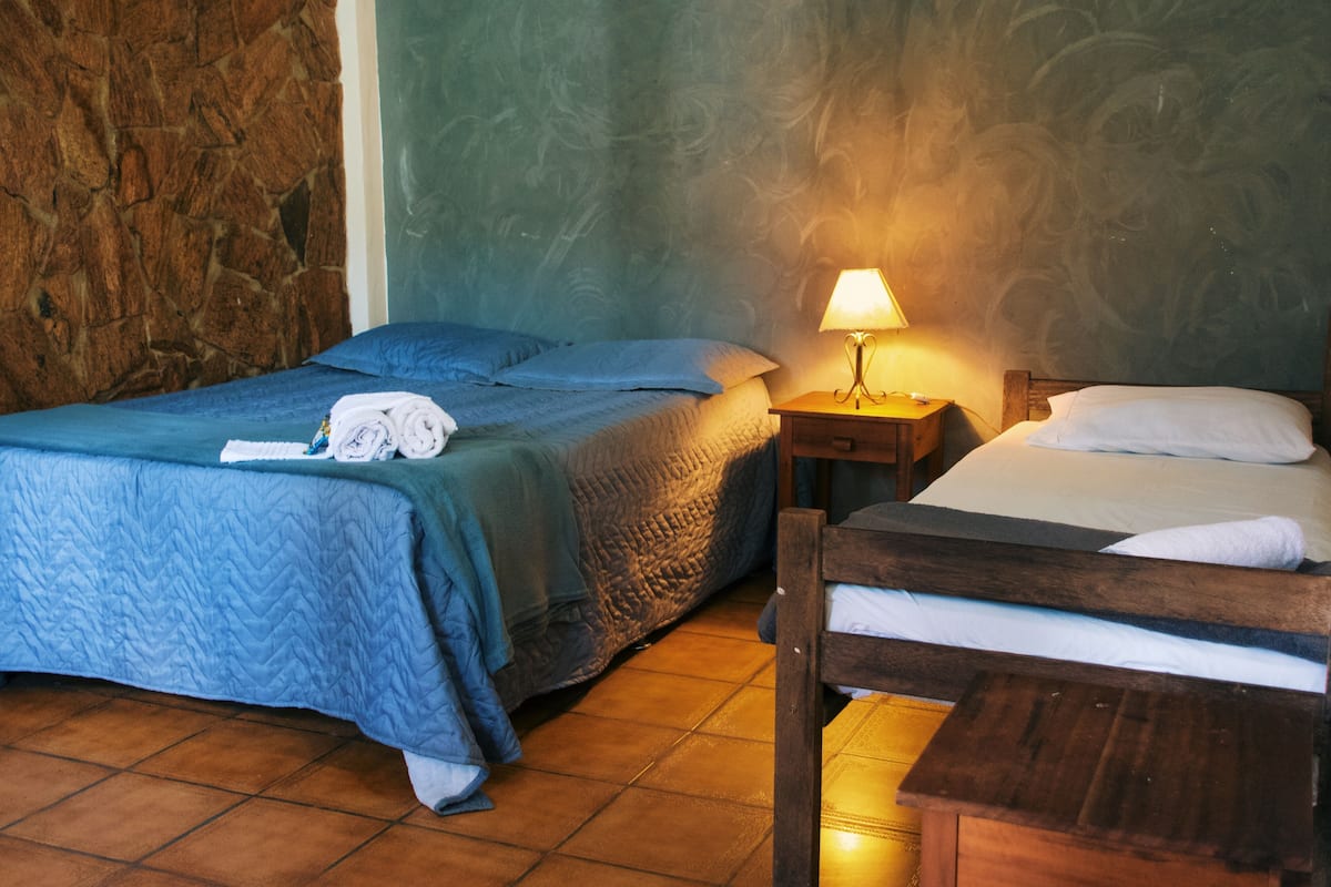 Una habitación con dos camas, una pared de piedra, suelo de madera y una mesita de noche con lámpara.