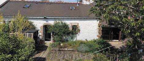 A white building with a tiled roof, a small garden area with plants, and a wooden structure to the left.
