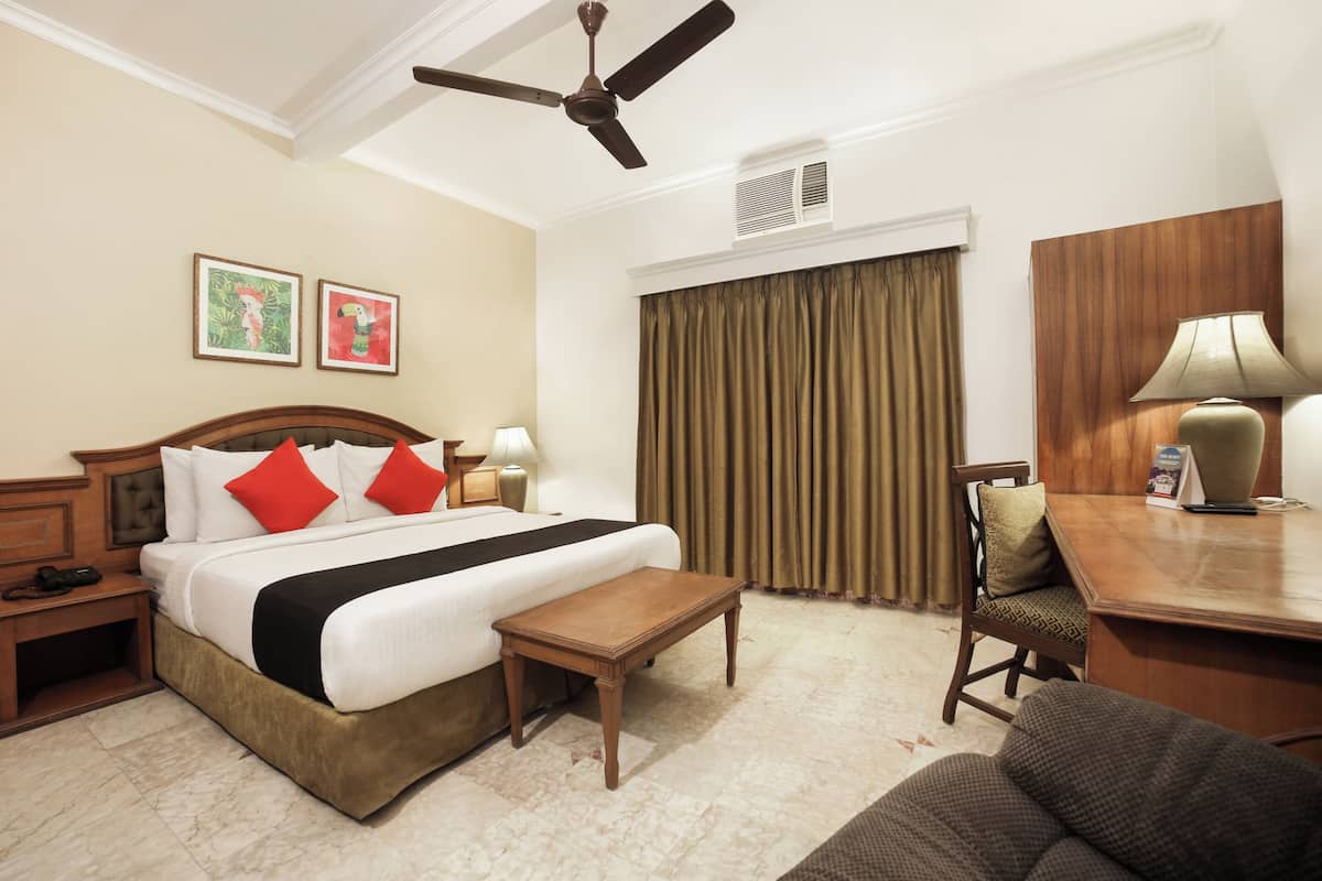 Quarto de hotel com cama, escrivaninha, cadeira, ventilador de teto e duas fotos emolduradas na parede.