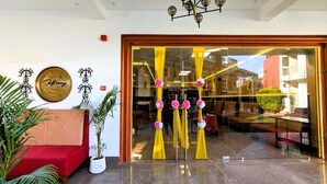 Entrada de hotel con un sofá rojo, plantas en macetas y elementos decorativos, incluyendo un letrero y cortinas.
