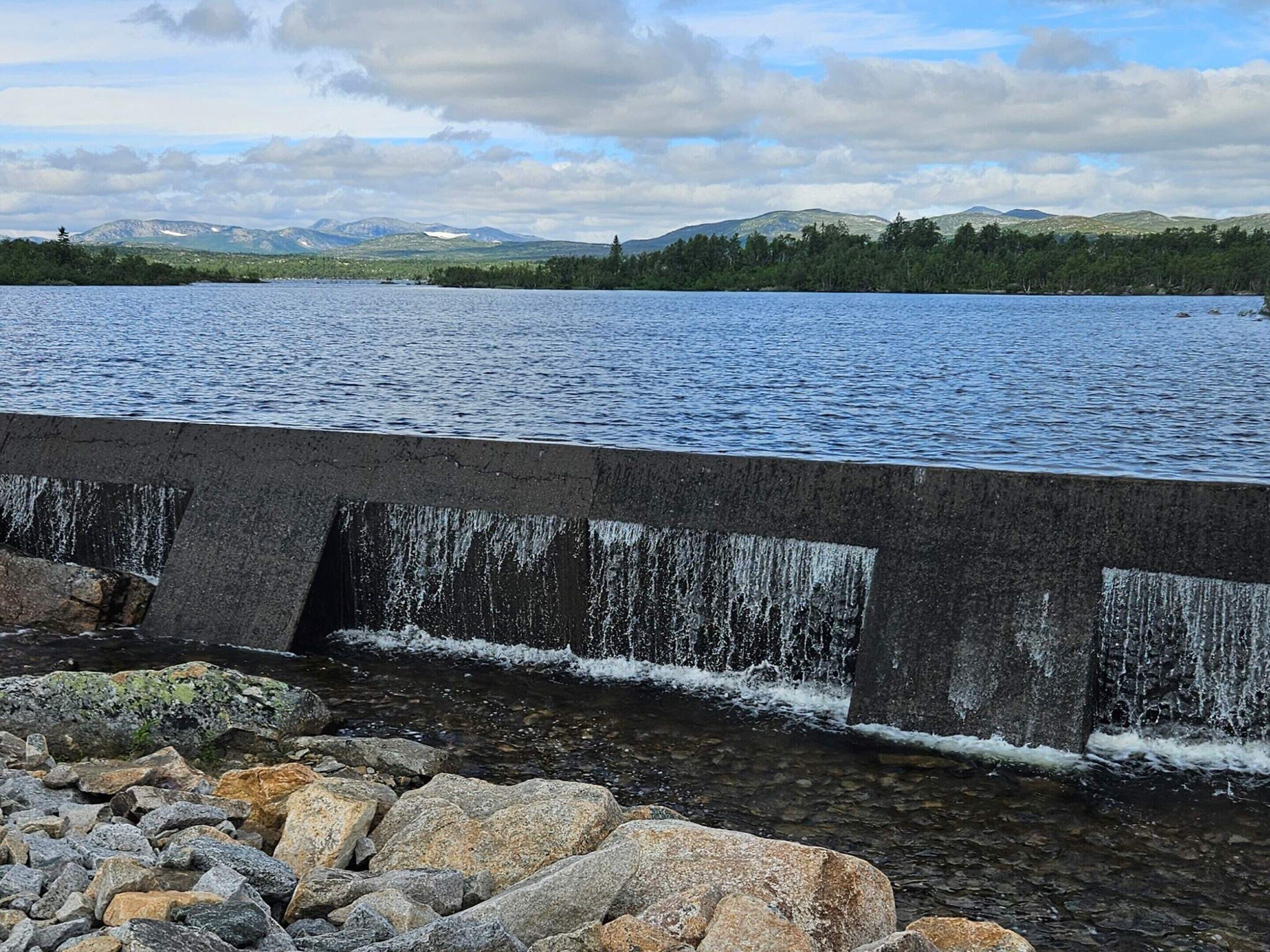 A concrete dam with water flowing over it, a rocky foreground, and a lake with mountains in the background.