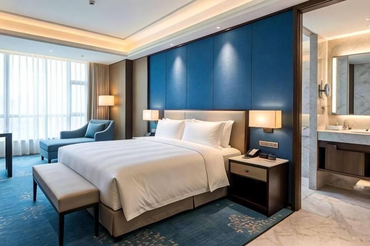Ett hotellrum med en stor säng, sängbord, ett skrivbord, en stol, en bänk, ett badrum med en spegel och ett fönster med gardiner.