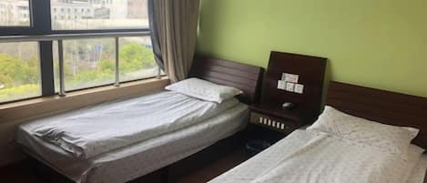 A hotel room with two single beds, a nightstand, and a window with a view of buildings and trees.