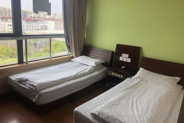 A hotel room with two single beds, a nightstand, and a window with a view of buildings and trees.