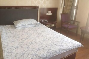 Un lit avec une couvre-lit à motifs floraux, une tête de lit en bois, une table de chevet avec une lampe, un fauteuil violet et une petite table.