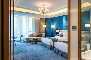 A hotel room with two beds, a sofa, a small table, a chandelier, a large window with curtains, and a bathroom with a mirror and sink.