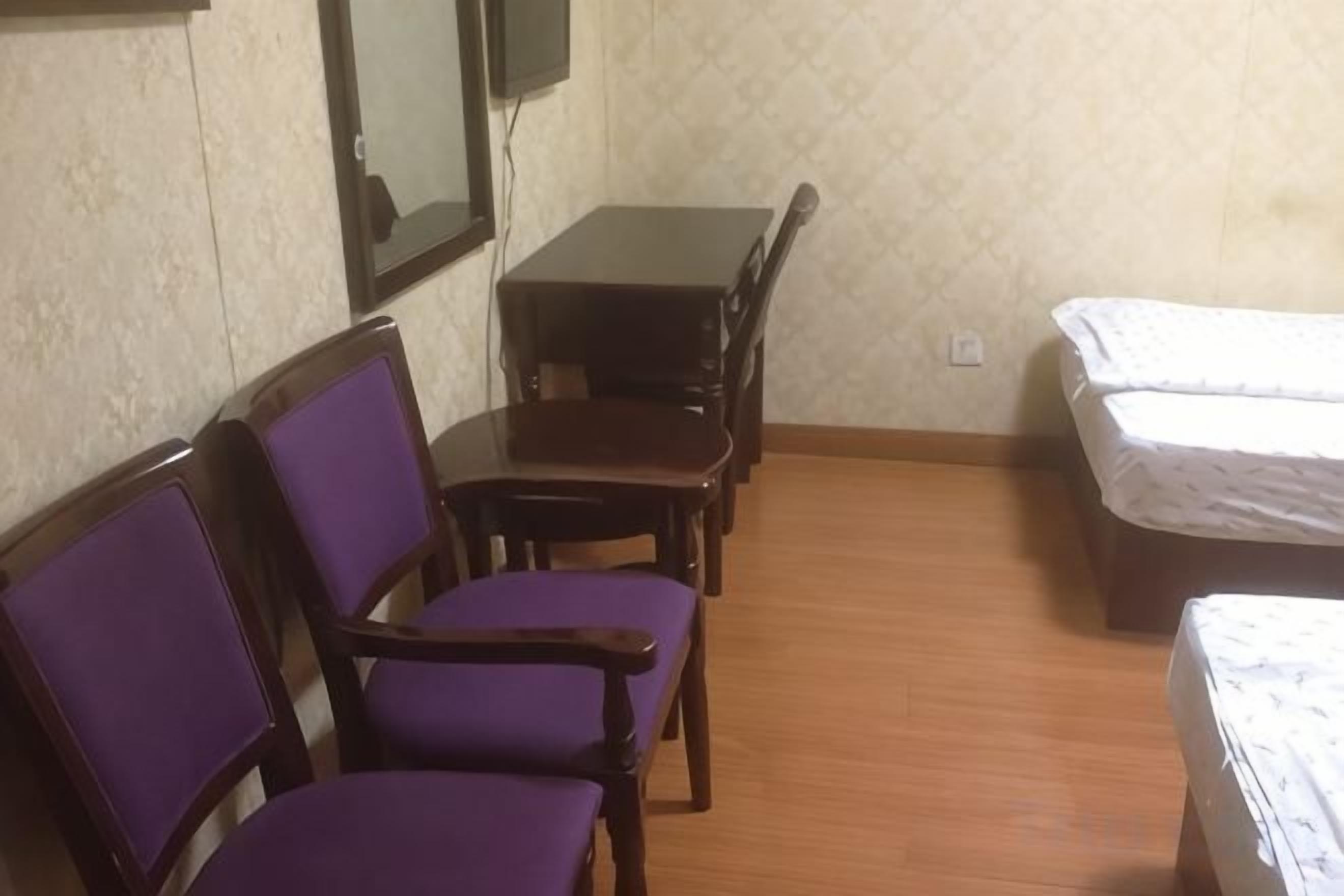 A room with a wooden floor, a bed with a white mattress, a small table with chairs, and a mirror on the wall.