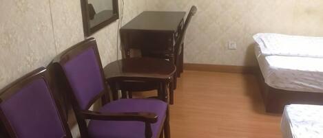 A room with a wooden floor, a bed with a white mattress, a small table with chairs, and a mirror on the wall.